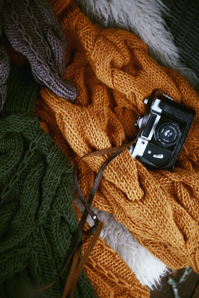 a grey, orange, and green knitted sweaters with an old camera on top of the orange sweater